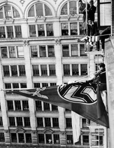 German consulate in San Francisco is today flying a Nazi Reich flag (with swastika); angry American crowd of 2000 has gathered outside & 2 sailors have climbed up 10 stories, trying to cut it down: Harold Sturtevant, a US Navy soldier on shore leave in San Francisco, has cut down the Nazi flag (after brief struggle with a German official)- to cheers of crowd.